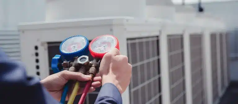 Hands holding HVAC manifold gauges near large outdoor air conditioning units, expertly conducting air conditioning maintenance in Nashville, TN.