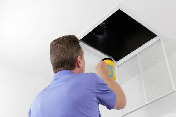 A person in a blue shirt is inspecting an open ceiling vent with a flashlight, looking into the dark interior space, potentially addressing air duct repair needs in Nashville, TN.