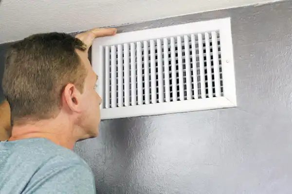 A person examines a rectangular air vent on a gray wall, considering the nuances of air duct installation in Nashville, TN.