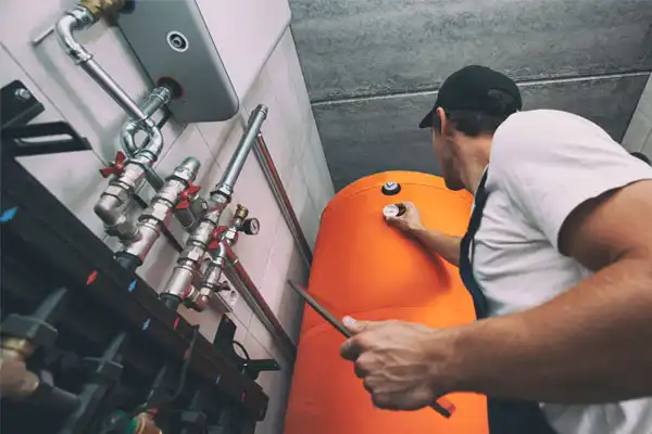 A technician in a cap inspects a large orange heating tank in a utility room in Nashville, surrounded by pipes and gauges, ensuring efficient heating repair.