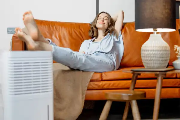 A woman relaxes on a leather sofa in Nashville, her feet resting on a blanket beside a trusty dehumidifier and a glowing table lamp, savoring the tranquil evening.