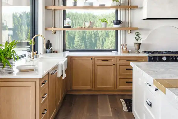 Modern kitchen in Jackson, TN, with light wood cabinets, white countertops, a window offering a forest view, gold faucet expertly installed after plumbing repair, open shelves adorned with plants, and a sleek black stove.
