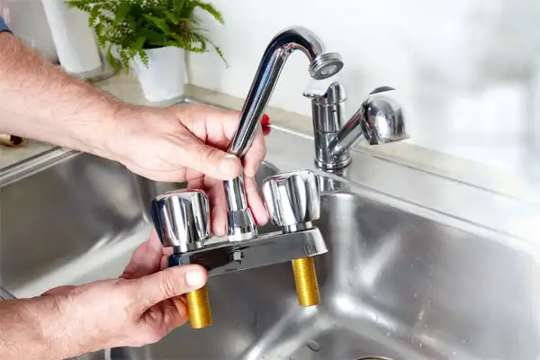 In Jackson, TN, a person showcases a detached chrome kitchen faucet with two handles above a stainless steel sink. A plant and soap dispenser add charm to the scene, hinting at an upcoming kitchen plumbing installation.