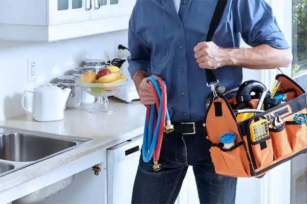 A person in a blue shirt, expertly handling plumbing hoses and a tool bag for kitchen plumbing repair in Jackson, TN, stands by a counter adorned with a fruit bowl and a white teapot.