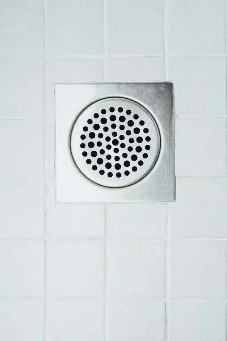 A square metal shower drain cover with circular holes is set in white tiled flooring, showcasing the precision and quality you expect from Rooter Services in Memphis.