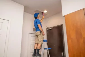 In a room with white walls, a person in blue shirt and shorts stands on a ladder to adjust a ceiling vent, ensuring top-notch air duct cleaning. This scene could easily be set in the vibrant city of Memphis, TN.