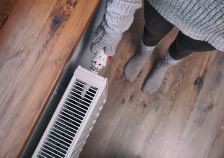 Person Wearing Knitted Gloves And Socks Adjusts A Radiator Thermostat In A Room With Wooden Flooring.